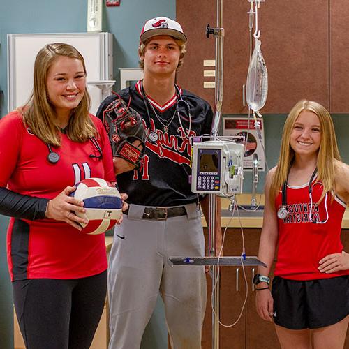 Three young adults in sports attire stand in a medical setting. One holds a volleyball, another a baseball glove, and the last one has an IV drip setup beside them.